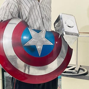 May include: A person holding a red, white and blue Captain America shield and a silver Thor's hammer. The shield has a large white star in the centre.