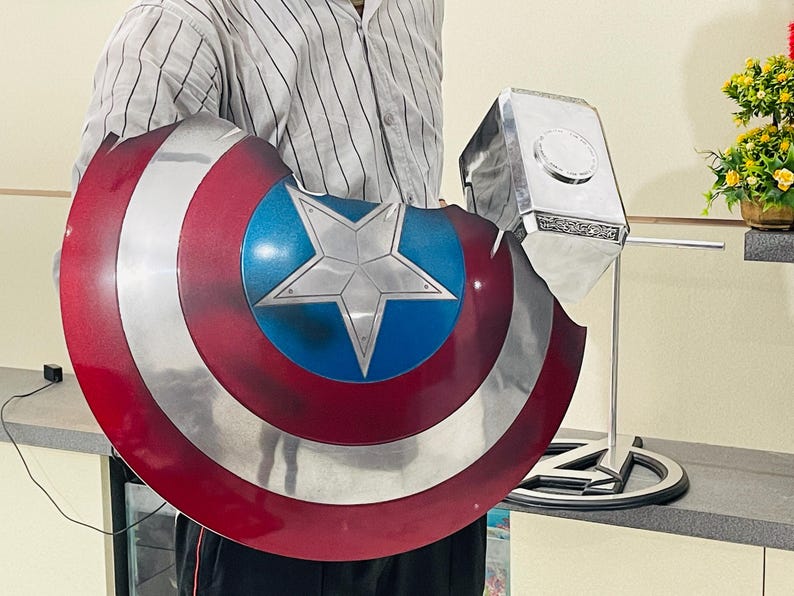 May include: A person holding a red, white and blue Captain America shield and a silver Thor's hammer. The shield has a large white star in the centre.