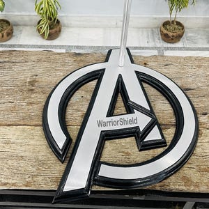 May include: A silver and black metal stand featuring the Avengers logo with the text "WarriorShield". The stand has a circular design with a large "A" in the center and a silver arrow pointing to the right.