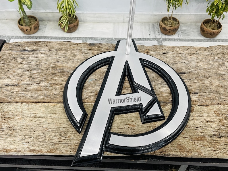 May include: A silver and black metal stand featuring the Avengers logo with the text "WarriorShield". The stand has a circular design with a large "A" in the center and a silver arrow pointing to the right.