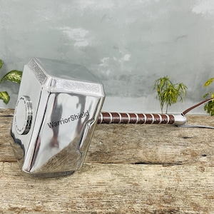 May include: A silver-colored hammer with a brown and silver handle and a leather strap. The hammer has the text "WarriorShield" on the side. The hammer is resting on a wooden surface.