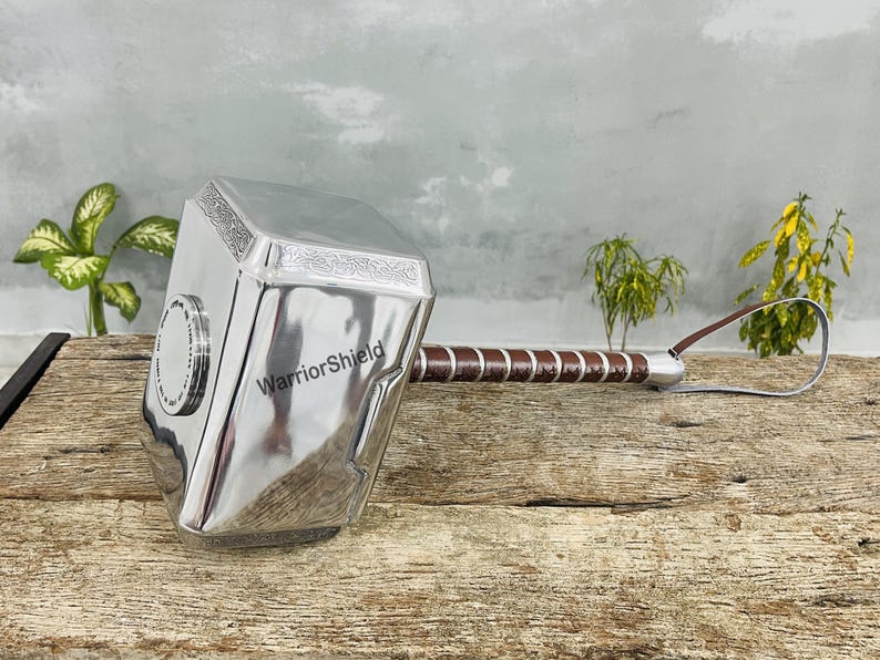 May include: A silver-colored hammer with a brown and silver handle and a leather strap. The hammer has the text "WarriorShield" on the side. The hammer is resting on a wooden surface.