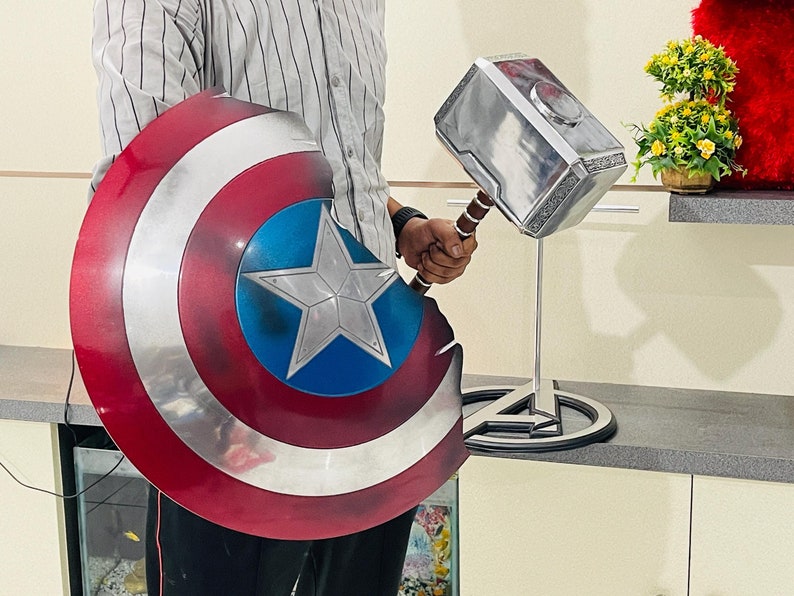 May include: A red, white and blue Captain America shield with a silver star in the centre. A silver Mjolnir hammer with a brown handle is on a stand with a black Avengers logo.