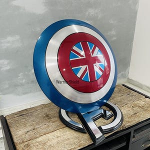 May include: A blue, red, and white Captain America shield with a Union Jack design in the center. The shield is on a black stand with the word "WarriorShield" printed on it.