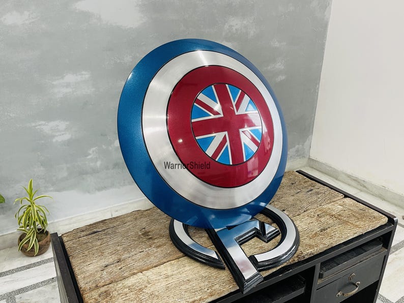 May include: A blue, red, and white Captain America shield with a Union Jack design in the center. The shield is on a black stand with the word "WarriorShield" printed on it.