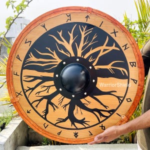 May include: A round wooden shield with a black and brown tree of life design. The shield has a black metal boss and is surrounded by a band of orange leather with runes.