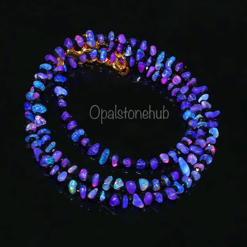 May include: A necklace composed of numerous small, irregularly shaped opals in shades of blue, purple, and pink. The opals are strung together, forming a circular shape. A gold-colored clasp secures the necklace. The background is black.