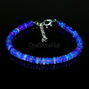 Op de afbeelding: Een blauwe opalen armband met een zilveren sluiting. De armband is gemaakt van kleine, ronde, gepolijste opalen kralen. De kralen hebben een levendige blauwe kleur met flitsen van andere kleuren. De tekst "OpalStoneHub" is zichtbaar.