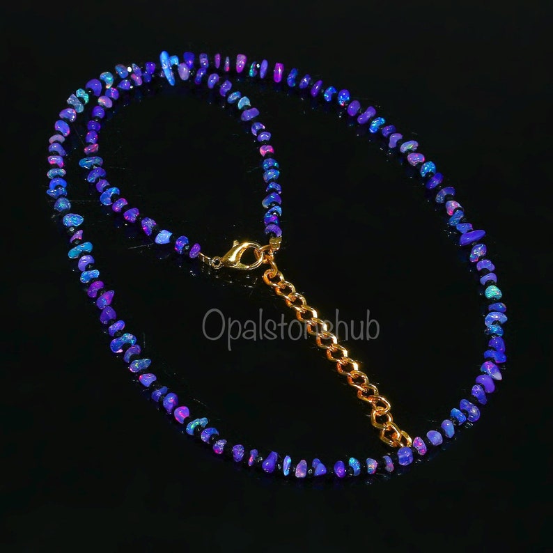 May include: A necklace featuring small, irregularly shaped blue and purple opal beads. The beads are strung together, creating a delicate and colorful piece. The necklace has a gold-colored clasp and chain extender.