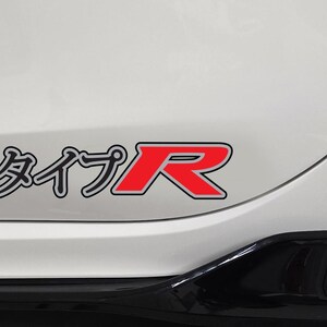 May include: A white car body panel featuring a decal with the Japanese katakana characters for Type R followed by a large, bold red letter R with a black outline and a white border, positioned above a black trim piece on the vehicle exterior.
