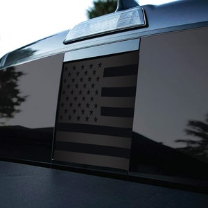 May include: A black and grey American flag decal for a vehicle window. The flag is displayed on a black truck window.