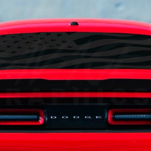 May include: A red Dodge Challenger with a black American flag decal on the rear window. The car has black taillights and a black spoiler.