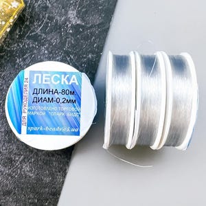 May include: Three spools of clear fishing line, each labeled "LESKA" with the text "ДЛИНА-80М ДИАМ-0,2ММ" in Cyrillic script. The spools are on a gray surface.