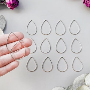 Puede incluir: Un conjunto de 12 anillos de salto de plata en forma de lágrima, dispuestos en una cuadrícula sobre una superficie gris. Los anillos de salto miden aproximadamente 2,5 cm de largo.
