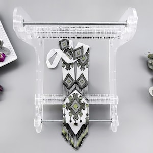 May include: A white and black beaded necklace with green accents hangs on a clear plastic loom. The necklace has a geometric pattern and a fringe.