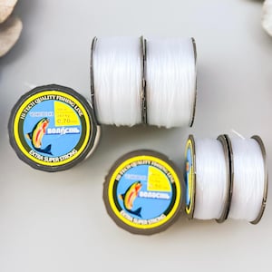 May include: Several spools of clear fishing line are arranged on a light gray surface. Each spool has a label with a blue and yellow design and the text "Extra Super Strong". Dried flowers and a decorative frame are also present.