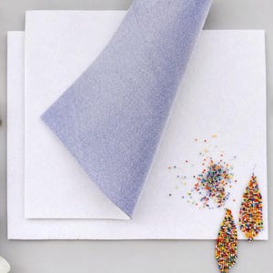 Könnte beinhalten: Zwei rechteckige Filzplatten, eine weiße und eine hellviolette, liegen übereinander. Neben den Filzplatten befinden sich zwei blattförmige Ohrringe aus kleinen, bunten Glasperlen sowie ein kleiner Haufen loser, farbenfroher Saatperlen.