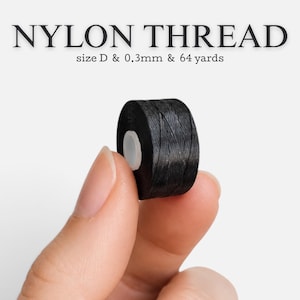 Könnte beinhalten: Eine kleine Spule aus schwarzem Nylongarn mit einem weißen Kern. Der Text NYLON THREAD size D & 0.3mm & 64 yards ist über der Spule aufgedruckt. Das Garn ist fest um die zylindrische Spule gewickelt und weist eine glatte, dunkle Textur auf.
