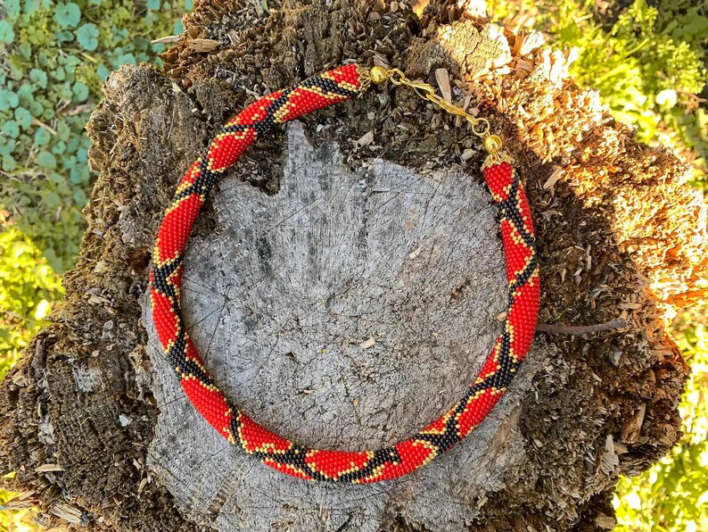 Puede incluir: Un collar de cuentas rojas, negras y doradas con forma de serpiente. El collar est&aacute; sobre un trozo de corteza de &aacute;rbol.