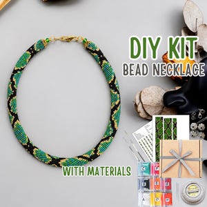 Könnte beinhalten: Ein DIY-Set für eine Perlenkette mit einer fertigen runden Halskette in einem schwarz-grün-goldenen Schlangenmuster. Das Set enthält eine gedruckte Anleitung, eine kleine Pappschachtel mit Schleife, beschriftete Perlenbeutel und Garn.