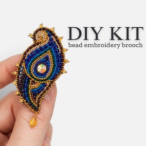May include: A DIY kit bead embroidery brooch featuring a paisley-shaped design in shades of blue, gold, and dark purple. The brooch is embellished with small beads and a central iridescent crystal, with a dangling amber-colored bead at the bottom.