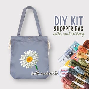 May include: DIY kit for a shopper bag with embroidery. The kit includes a gray canvas tote bag, embroidery floss, and beads. The bag features a white daisy with a yellow center.