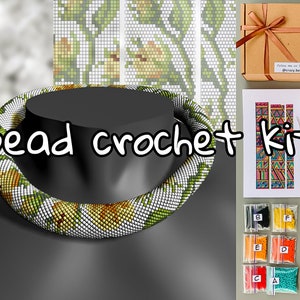 May include: A bead crochet kit featuring a white and green beaded necklace, a pattern sheet, and a selection of beads in small bags. The text "Bead crochet kit" is written in black marker on a black surface.