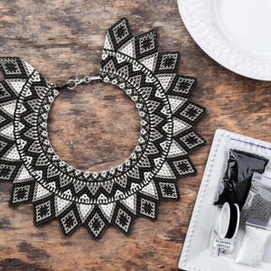 May include: A black and white beaded necklace with a geometric pattern. The necklace is in the shape of a collar and has a large, circular design in the center. The necklace is lying on a wooden surface.
