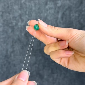 May include: A close-up of a green bead on a string held between two hands. The bead is round and has a marbled appearance. The string is white and thin. The background is a dark gray color.