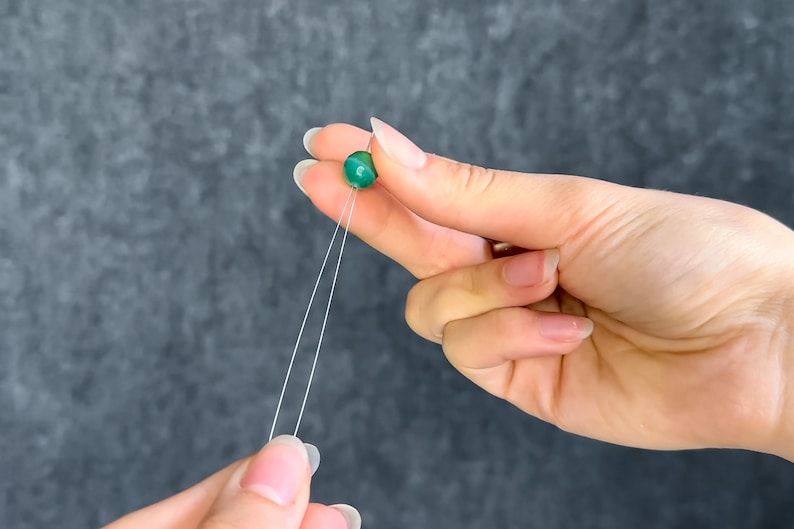 May include: A close-up of a green bead on a string held between two hands. The bead is round and has a marbled appearance. The string is white and thin. The background is a dark gray color.