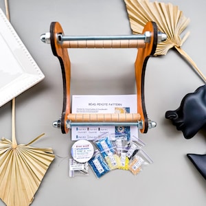 May include: A wooden bead loom with a metal rod and wooden dowels. The loom is surrounded by small bags of colorful beads, a spool of thread, and a bead weaving pattern.