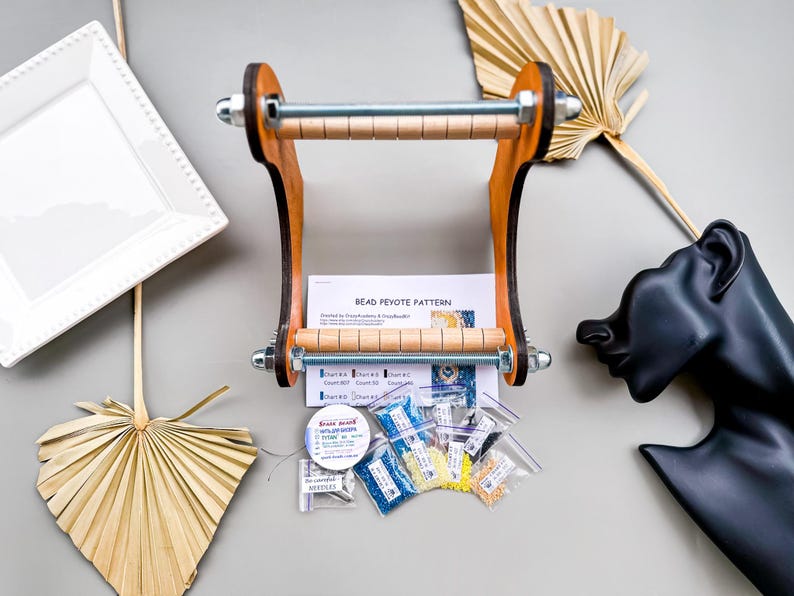May include: A wooden bead loom with a metal rod and wooden dowels. The loom is surrounded by small bags of colorful beads, a spool of thread, and a bead weaving pattern.