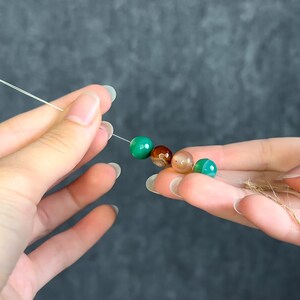 May include: A string of colorful gemstone beads, including green, brown, and tan, held between two hands. The beads are strung on a thin, clear cord, and a piece of twine is held in the other hand. The background is a solid, dark gray.