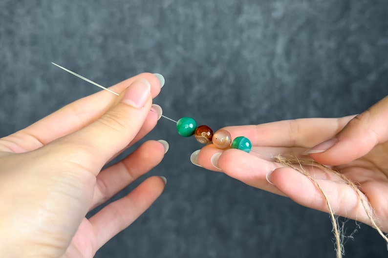 May include: A string of colorful gemstone beads, including green, brown, and tan, held between two hands. The beads are strung on a thin, clear cord, and a piece of twine is held in the other hand. The background is a solid, dark gray.