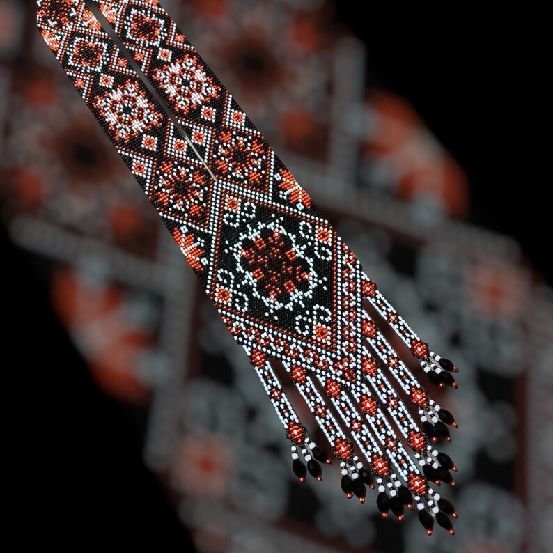 Op de afbeelding: Een paar lange, kralenkettingen met zwarte, witte en oranje geometrische patronen. De kettingen hebben franjes aan de onderkant.