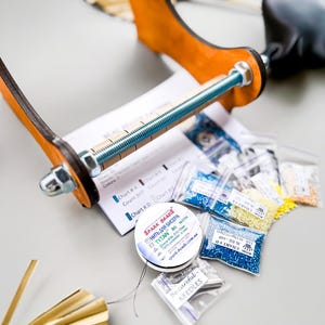 May include: A close-up of a bead loom with a wooden frame and a metal screw. There are several small bags of colorful beads, a spool of thread, and a package of needles.
