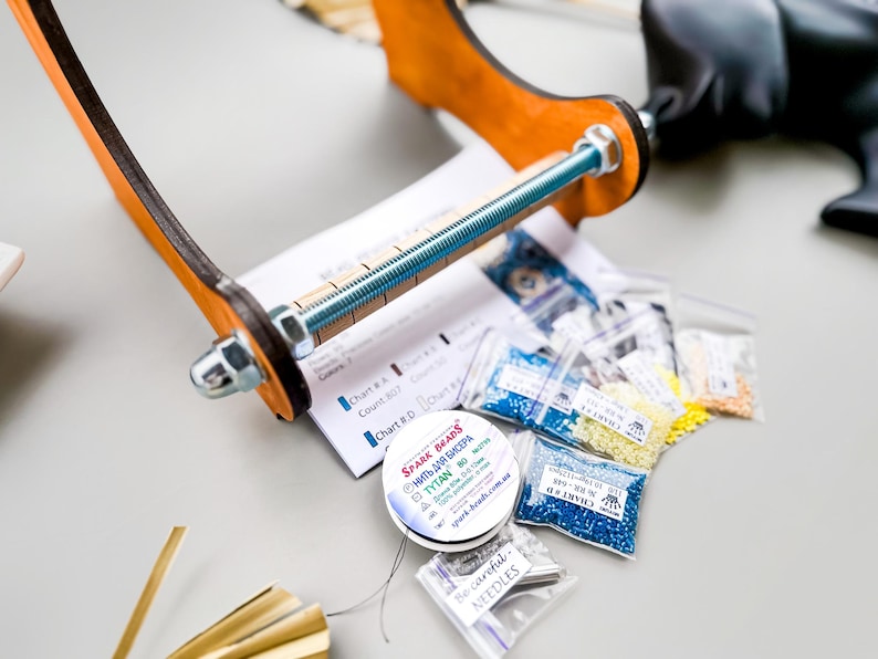 May include: A close-up of a bead loom with a wooden frame and a metal screw. There are several small bags of colorful beads, a spool of thread, and a package of needles.