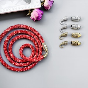 Peut inclure: Un collier de perles rouges avec des embouts en forme de serpent. Le collier est enroul&eacute; et pos&eacute; &agrave; plat sur une surface grise. Il y a aussi plusieurs embouts en forme de serpent argent&eacute;s et dor&eacute;s sur la surface.