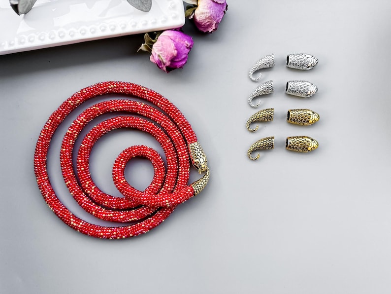Peut inclure: Un collier de perles rouges avec des embouts en forme de serpent. Le collier est enroul&eacute; et pos&eacute; &agrave; plat sur une surface grise. Il y a aussi plusieurs embouts en forme de serpent argent&eacute;s et dor&eacute;s sur la surface.