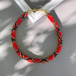 Puede incluir: Un collar de cuentas rojas, negras y doradas con un patr&oacute;n de piel de serpiente.