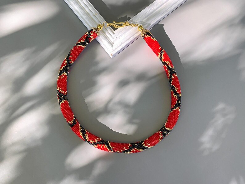 Puede incluir: Un collar de cuentas rojas, negras y doradas con un patr&oacute;n de piel de serpiente.