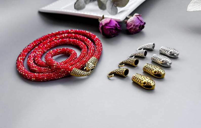 Peut inclure: Un collier de perles rouges avec des fermoirs en forme de serpent. Le collier est enroul&eacute; sur une surface grise. Il y a aussi plusieurs fermoirs en forme de serpent argent&eacute;s et dor&eacute;s sur la surface.