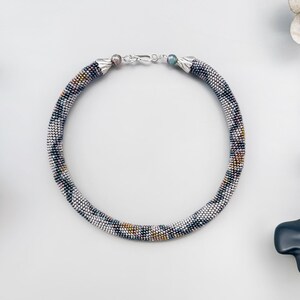 May include: A beaded necklace with a silver clasp. The necklace is a tube shape, with a pattern of white, black, and gold beads. The necklace is on a white surface with decorative elements.