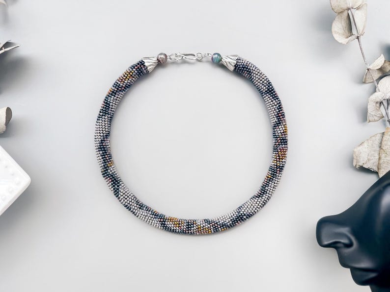 May include: A beaded necklace with a silver clasp. The necklace is a tube shape, with a pattern of white, black, and gold beads. The necklace is on a white surface with decorative elements.
