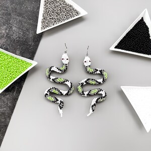 May include: A pair of beaded snake earrings with green, white, and black accents. The earrings are displayed on a white surface with small bowls of beads.