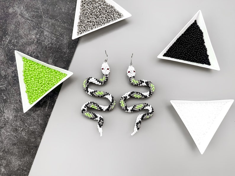 May include: A pair of beaded snake earrings with green, white, and black accents. The earrings are displayed on a white surface with small bowls of beads.