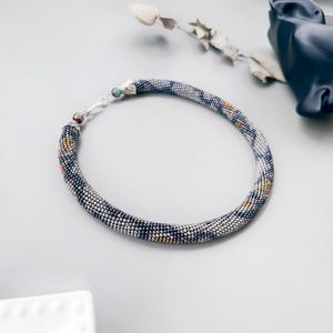 May include: A beaded necklace with a silver clasp. The necklace is a tube shape, with a pattern of small beads in shades of blue, white, yellow, and black. The necklace is on a light gray surface.