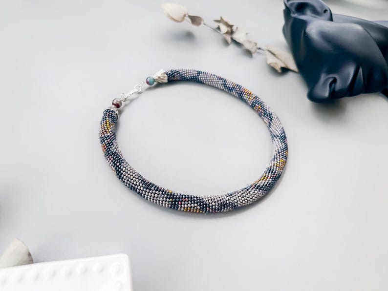 May include: A beaded necklace with a silver clasp. The necklace is a tube shape, with a pattern of small beads in shades of blue, white, yellow, and black. The necklace is on a light gray surface.