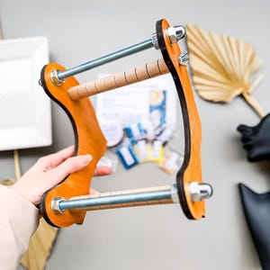 May include: A wooden loom with two metal rods and a wooden frame. The loom is held in a person's hand.