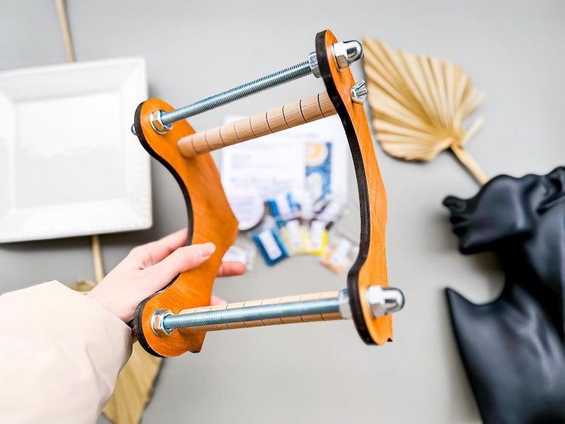 May include: A wooden loom with two metal rods and a wooden frame. The loom is held in a person's hand.